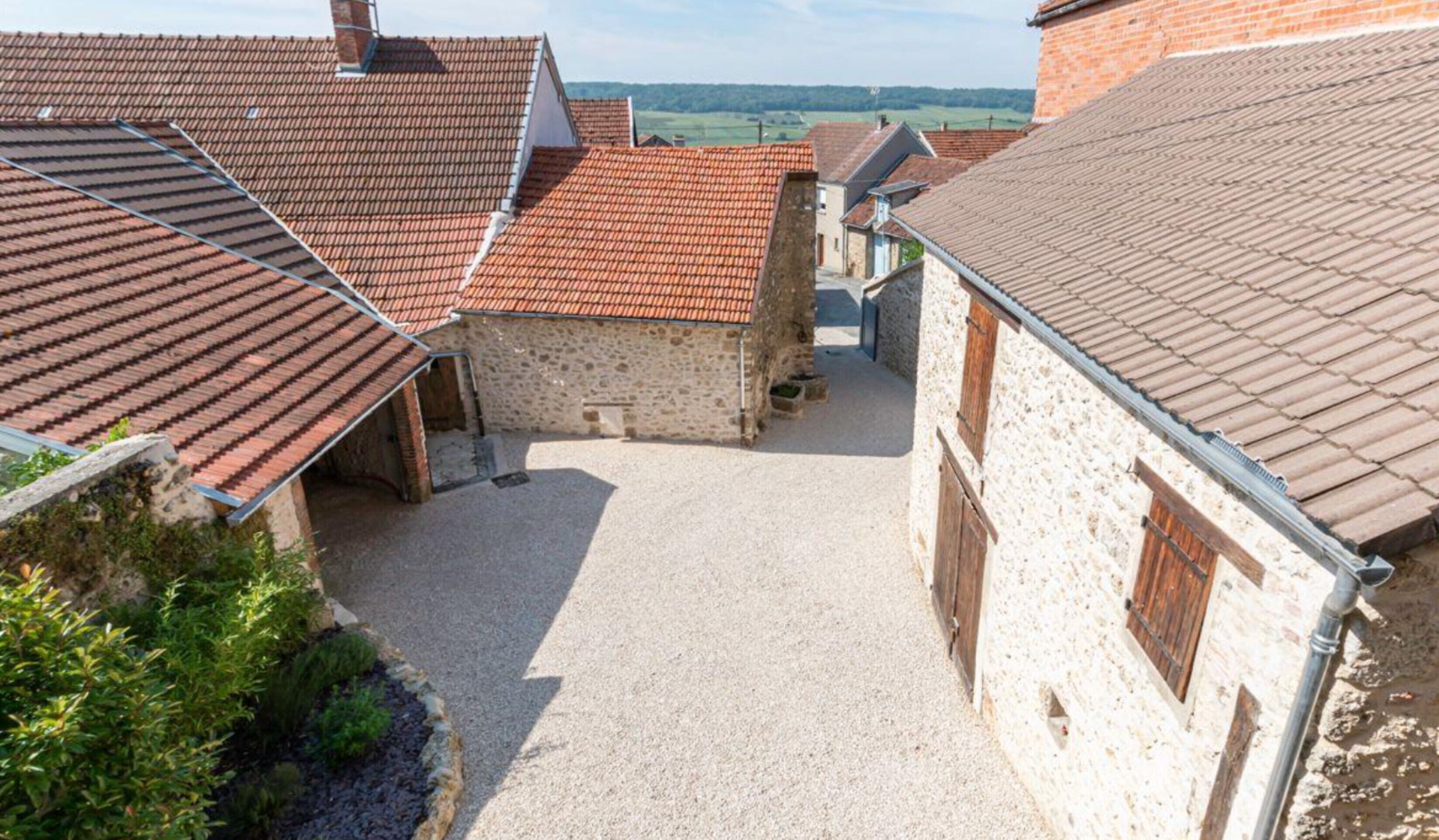 Une cour bordée de bâtiments en pierre, dotée de toits couverts de tuiles rouges et d’un chemin de gravier.