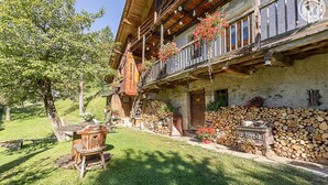 A traditional wooden chalet with a balcony, a stone wall, and a log stack.