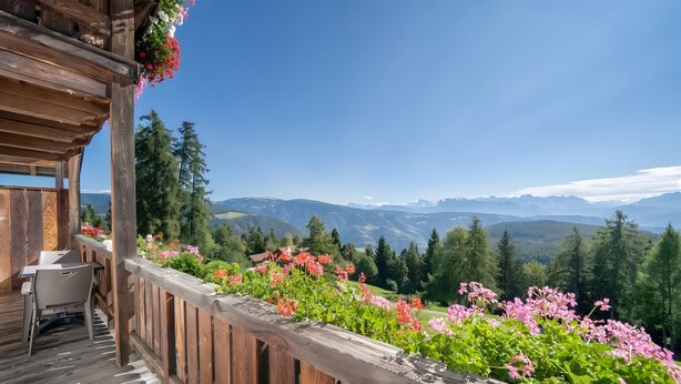 Una terraza de madera con mesa y sillas, rodeada de plantas en flor, con vistas a un paisaje montañoso.