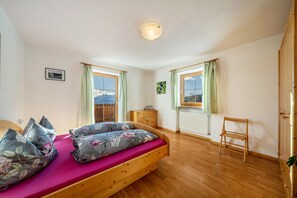 Quarto com cama e cadeira de madeira, radiador e janela com vista para as montanhas.