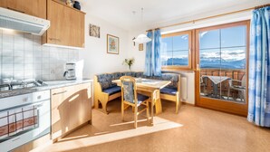 Una cocina con estufa, horno y cafetera, un comedor con mesa y sillas, y un amplio ventanal con vistas a las montañas.