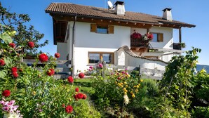 Una casa de dos pisos con techo de tejas, un balcón con flores y un jardín con diversas plantas y flores.