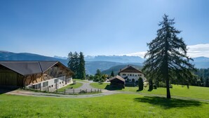 Un paysage rural avec une grange, un bâtiment blanc doté d’un toit en pente et un grand arbre à feuilles persistantes.