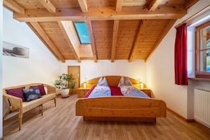 A bedroom with a wooden ceiling, a bed with pillows, a wicker sofa, a potted plant, and a window with curtains.