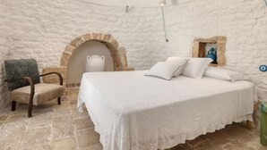 A room with a stone wall, a bed with white linens, a chair, and a small table with a pitcher. - Villa 'Borgo Trulli 1817' with Private Pool, Wi-Fi and Air Conditioning (Putignano)