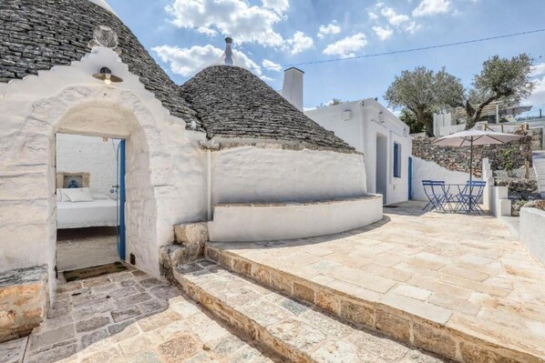 Une maison traditionnelle en pierre, avec une façade blanche, un escalier en pierre menant à une porte, et un petit espace extérieur aménagé avec une table et des chaises.