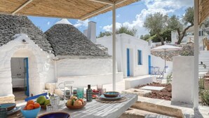 An outdoor dining area with a table set for two, surrounded by white buildings with blue doors, under a thatched roof. - Villa 'Borgo Trulli 1817' with Private Pool, Wi-Fi and Air Conditioning (Putignano)