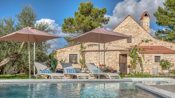 Un edificio de piedra con piscina, sillas de descanso y sombrillas.