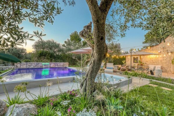 A swimming pool surrounded by greenery, a stone building, and outdoor seating.