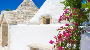 A white-washed building with a conical roof, a stone structure, and a potted plant with pink flowers. - Villa 'Trulli Life' with Private Pool, Wi-Fi and Air Conditioning (Putignano)