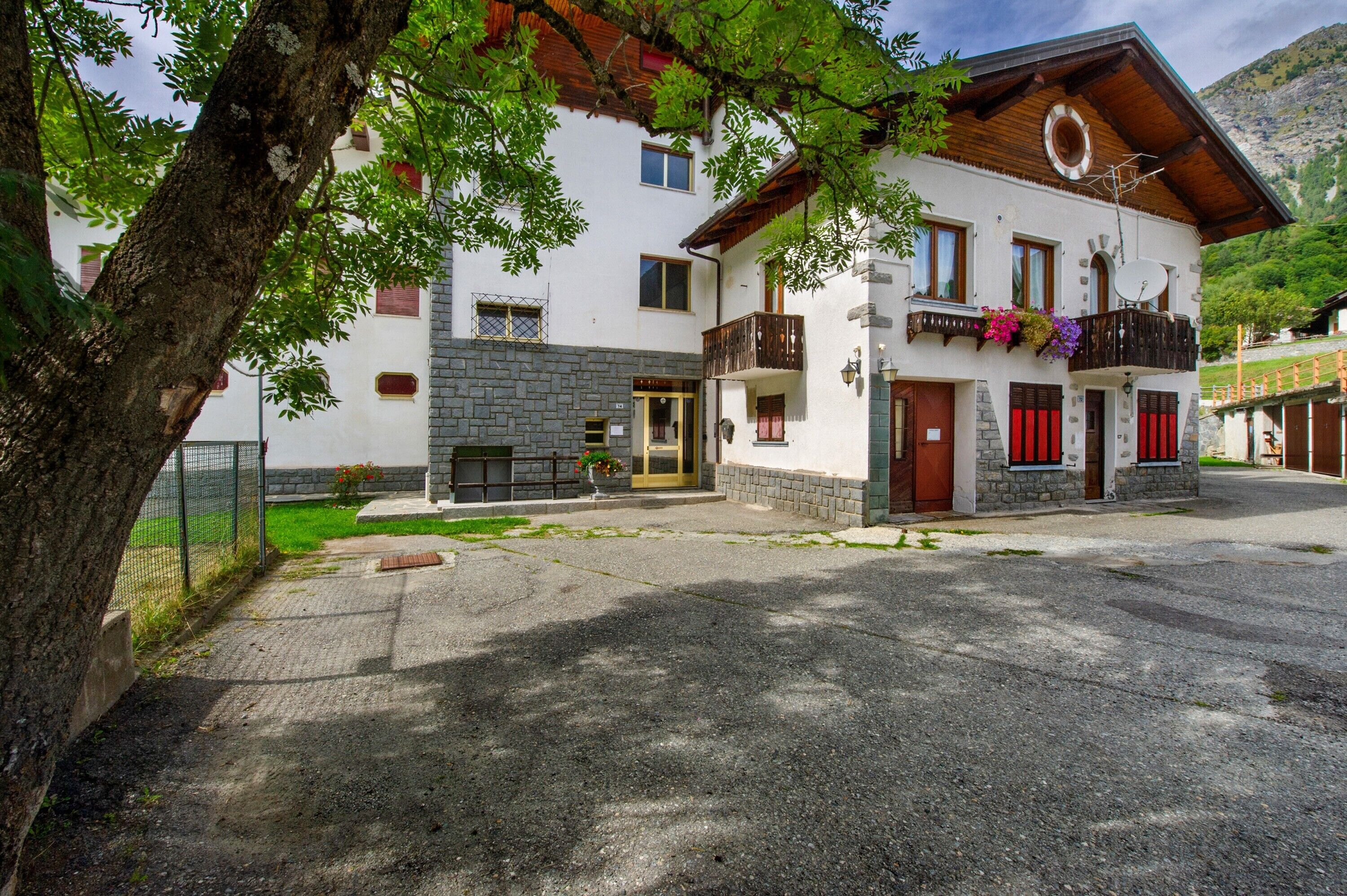 Un immeuble de deux étages avec une façade en pierre, un balcon agrémenté de jardinières et un paysage montagneux en arrière-plan.