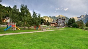 Un parque infantil con toboganes y columpios, un campo cubierto de césped y edificios con balcones en una zona montañosa.