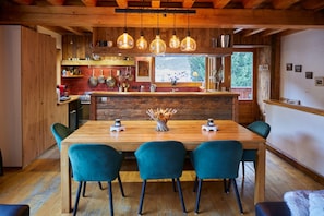 A wooden dining area with a long table and four chairs, a kitchen with hanging pots, and a window with a view of greenery.