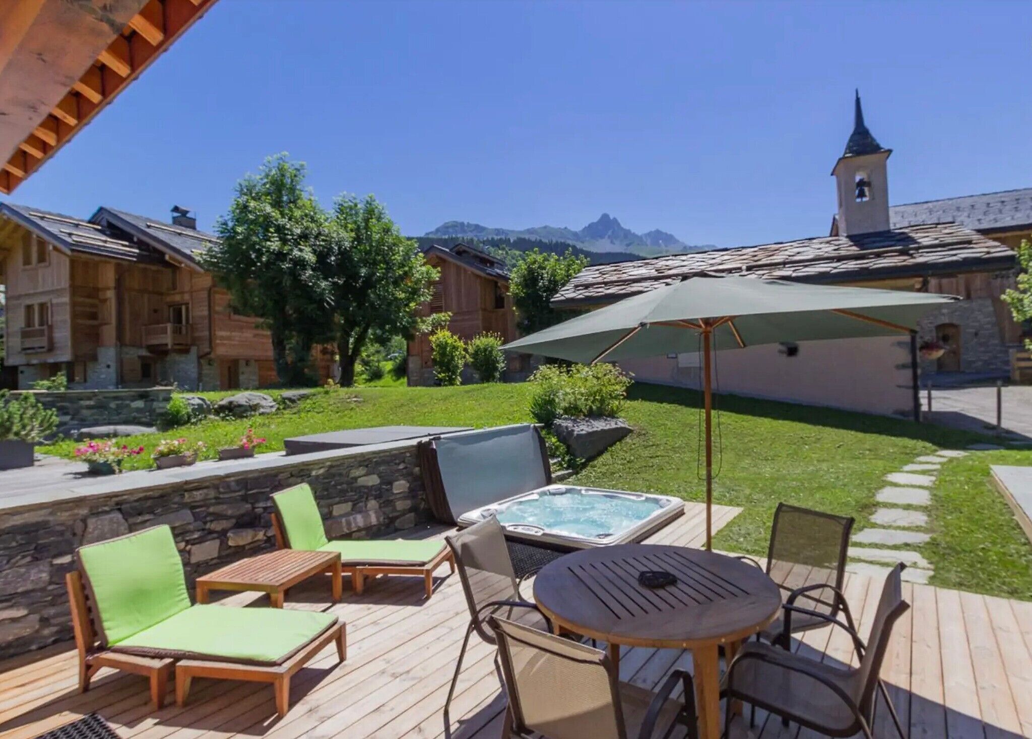 A wooden deck with lounge chairs, a hot tub, and a view of mountains and a church tower.
