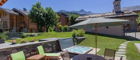 A wooden deck with lounge chairs, a hot tub, and a view of mountains and a church tower.