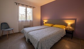 A bedroom with two beds, a chair, a window with curtains, and a nightstand with lamps.