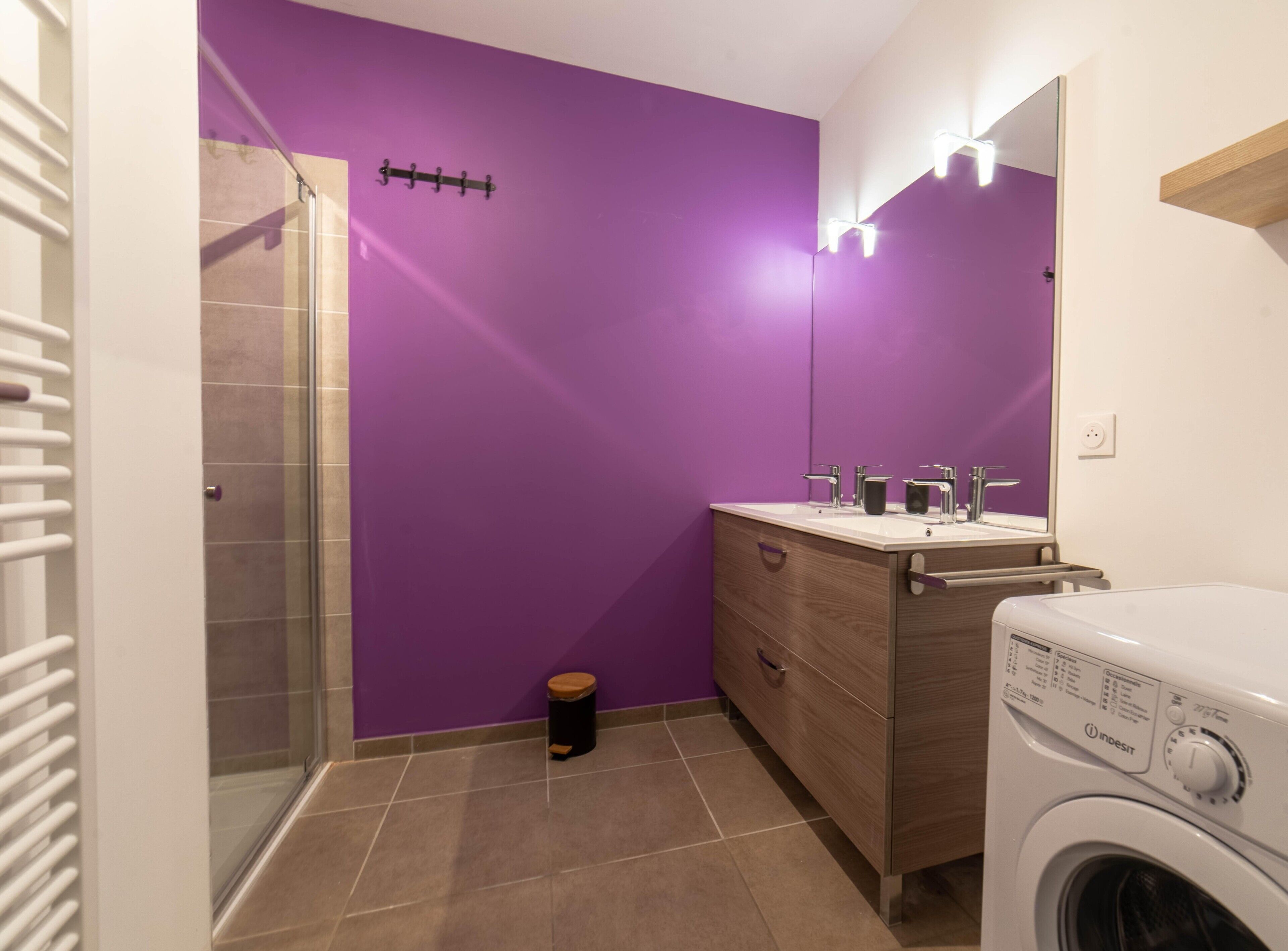 Un baño con una pared morada, una cabina de ducha de vidrio, un tocador de madera con espejo y una lavadora de carga frontal.