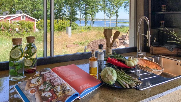 A kitchen counter with a cookbook, a bottle of oil, a basket of vegetables, and a view of a lake and trees.