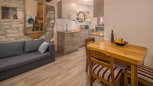 A cozy living room with a grey sofa, a wooden dining table with chairs, a stone wall, and a kitchen area with wooden cabinets and a microwave.