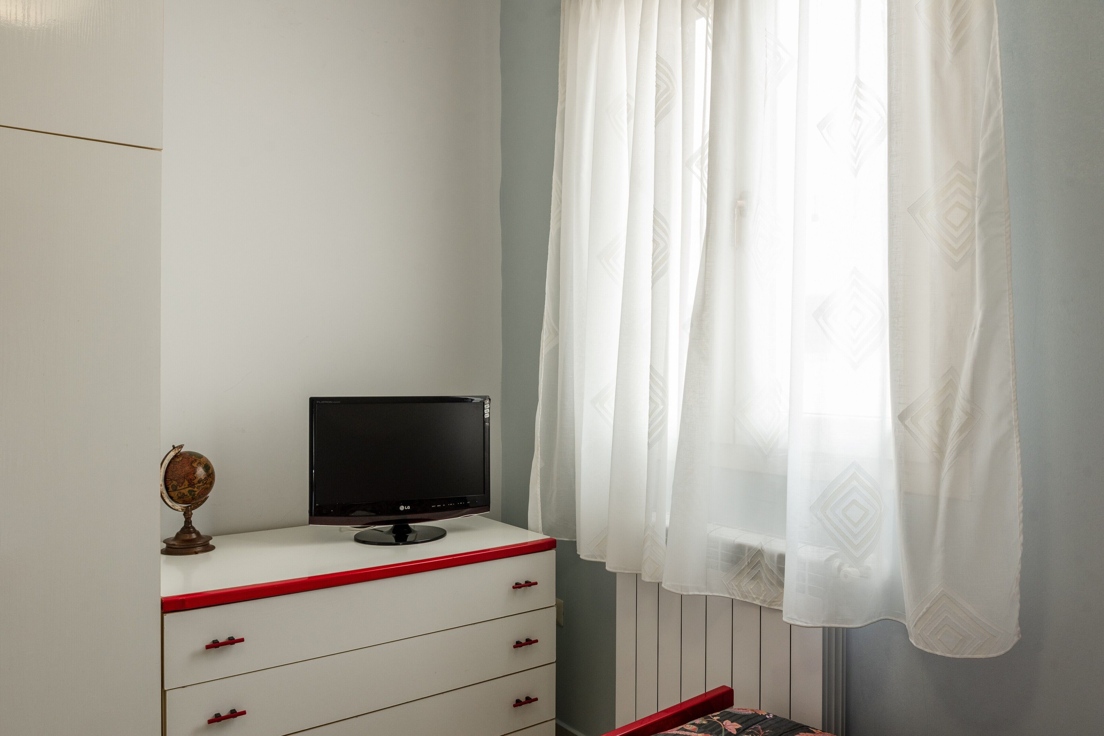 A room with a white desk, a closed black monitor, a globe, and a window with sheer white curtains.