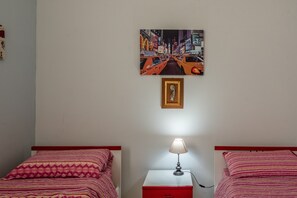 Une chambre d’hôtel avec deux lits, une table de chevet avec une lampe, un tableau encadré au mur et un tiroir rouge.