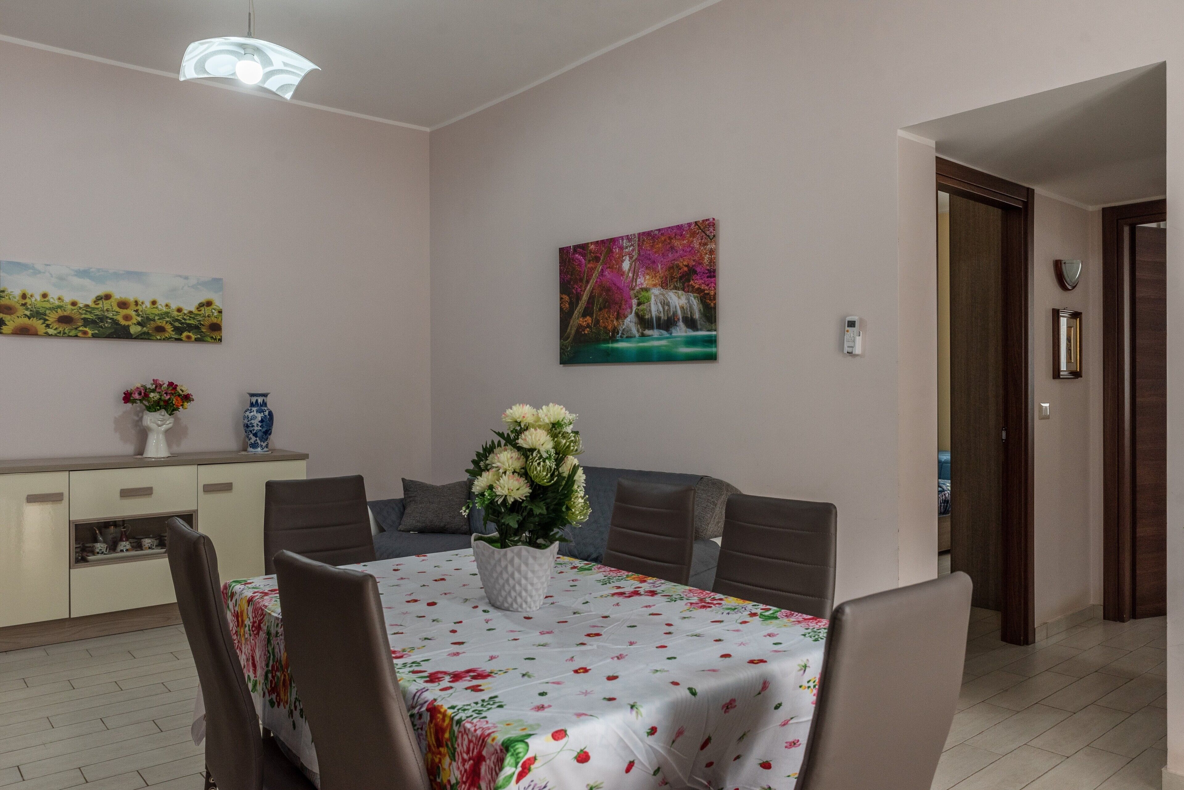 Uma sala de jantar com uma mesa posta para quatro pessoas, um vaso com flores e um quadro na parede.