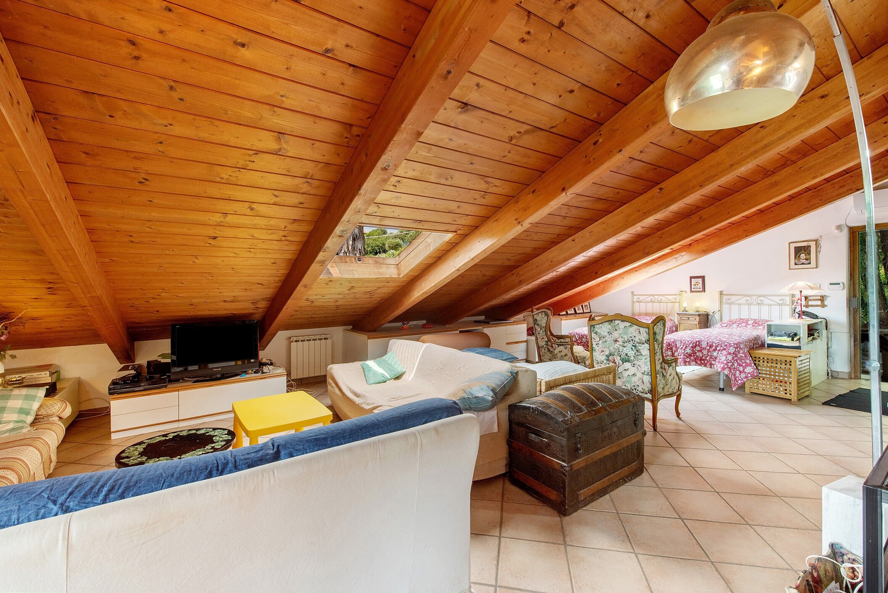 A cozy attic bedroom with a wooden ceiling, a bed, a sofa, a television, and a window overlooking greenery.