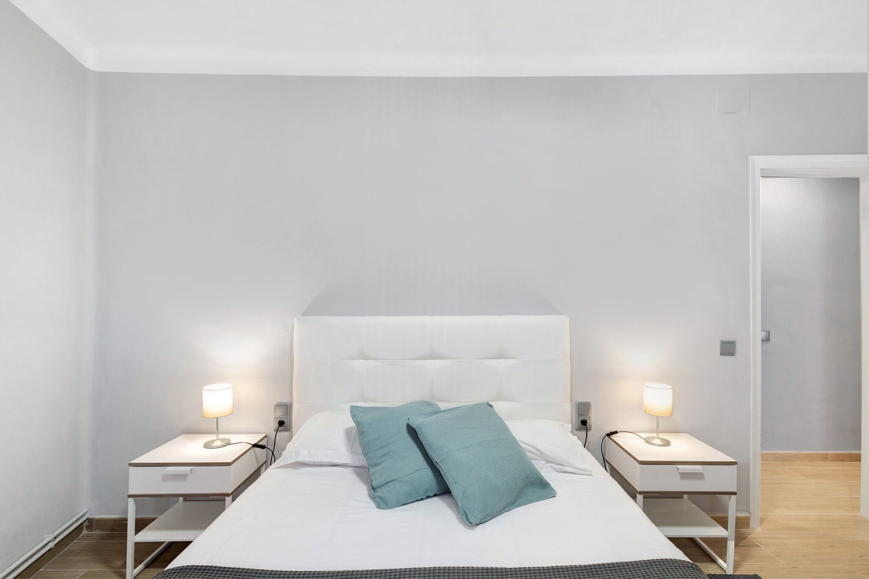 Una camera da letto con un letto, due comodini con lampade, una testiera bianca e una porta sulla destra.