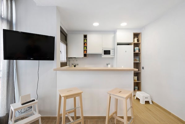 A modern kitchen with a flat-screen TV, a microwave, and a small refrigerator. There are two wooden stools and a white step stool.