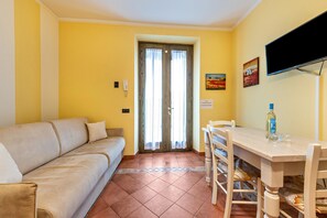Una habitación con un sofá color beige, una mesa de comedor de madera con sillas, un televisor en la pared y una puerta con cortinas.