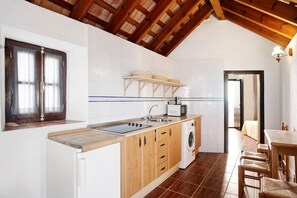 Una cocina con armarios de madera, lavadora, microondas y ventana con cortinas.