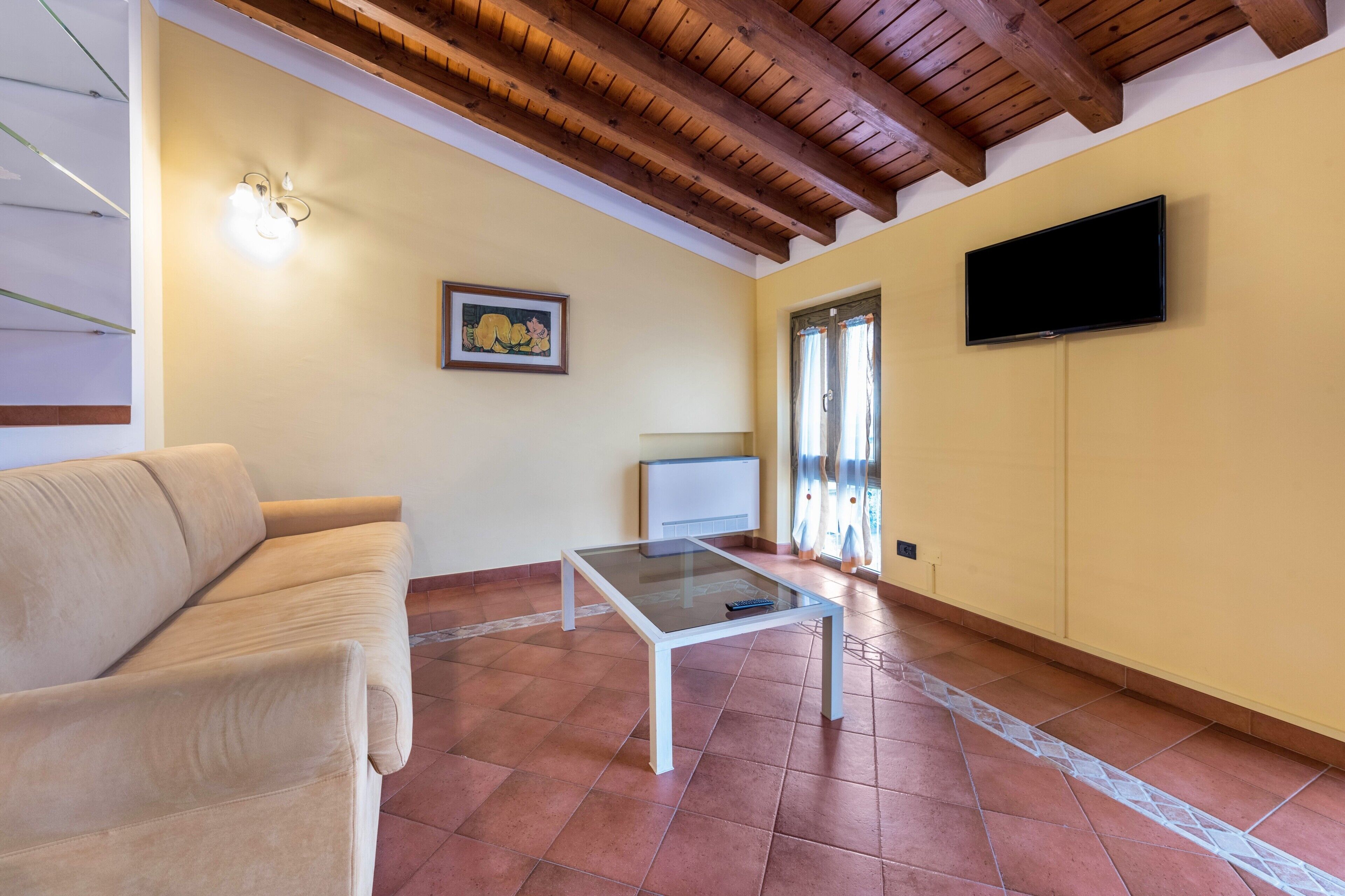 A living room with a sofa, a glass-top coffee table, a flat-screen TV, a painting, and a tiled floor.