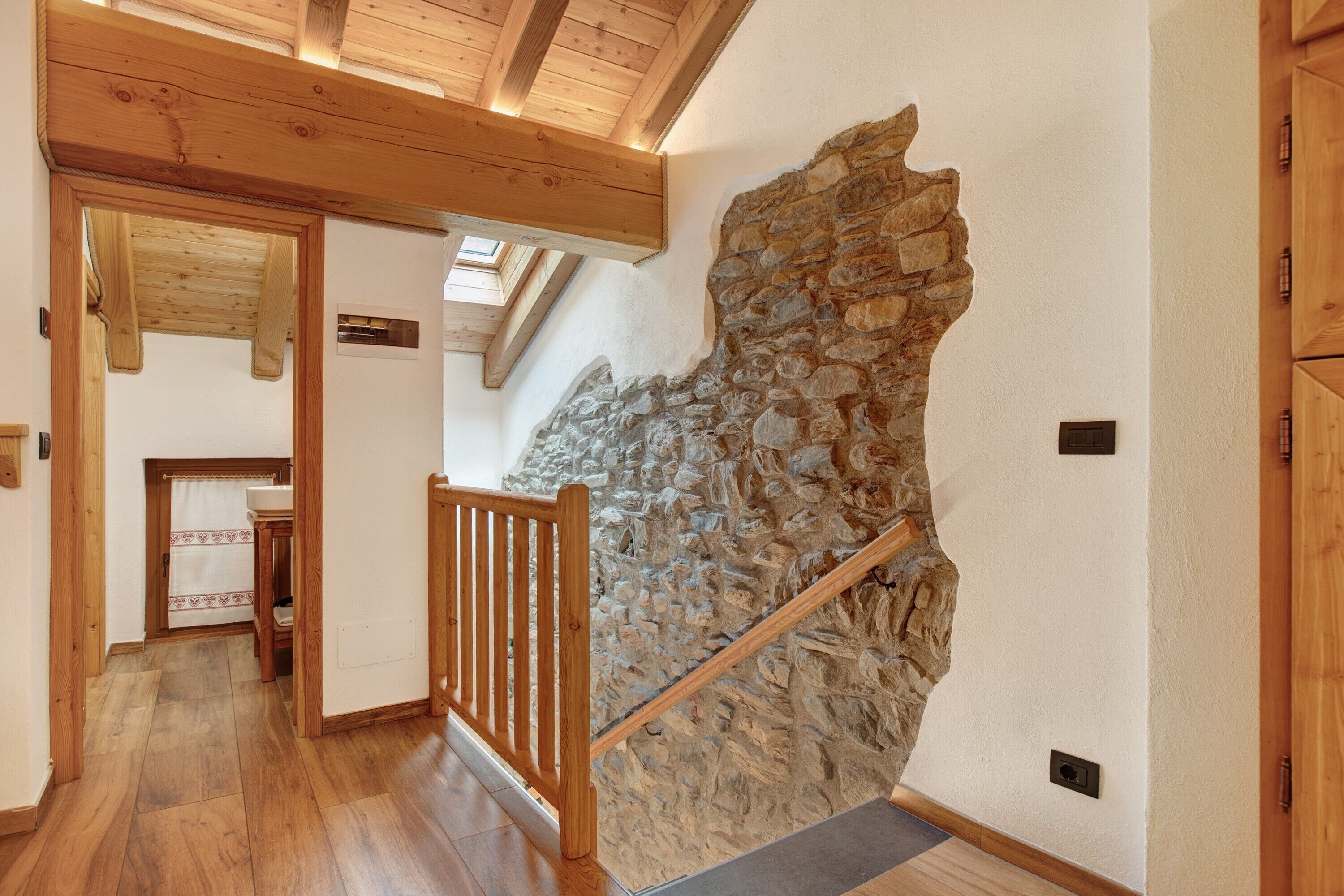 A wooden staircase with a stone wall, a wooden railing, and a view of a kitchen area.