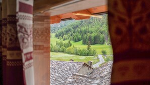A view through a window with a patterned curtain, a wooden ceiling, and a landscape with trees and a road. - Studio 'Aux Pieds Du Malatra' 1 with Mountain View (Saint Rhemy en Bosses)