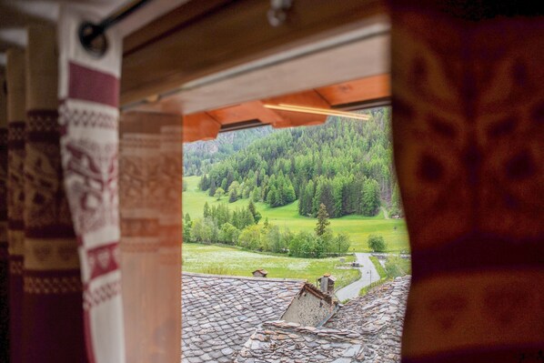 A view through a window with a patterned curtain, a wooden ceiling, and a landscape with trees and a road.
