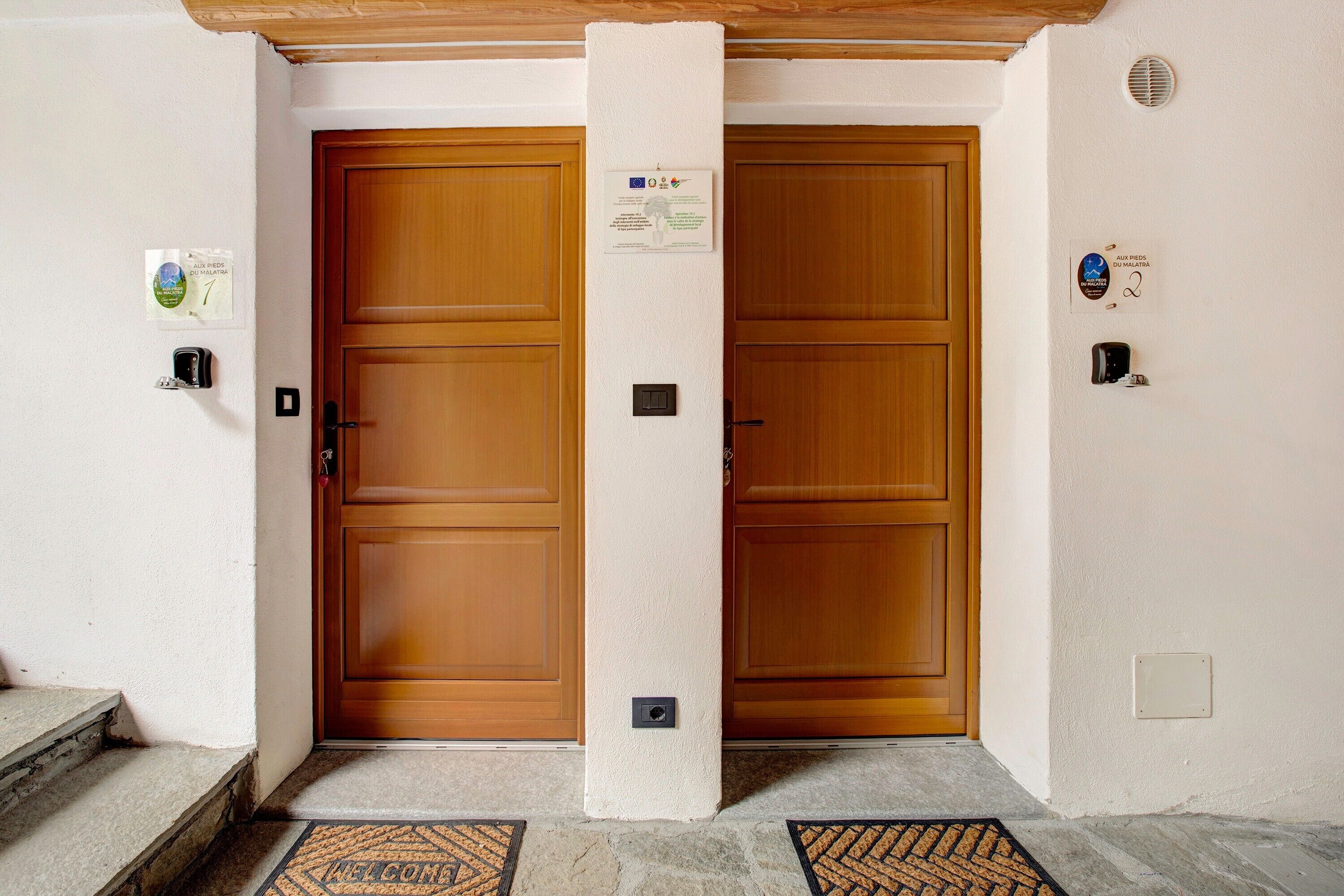 L'ingresso di un hotel con due porte di legno, un tappetino d'entrata e una piccola insegna sulla parete.