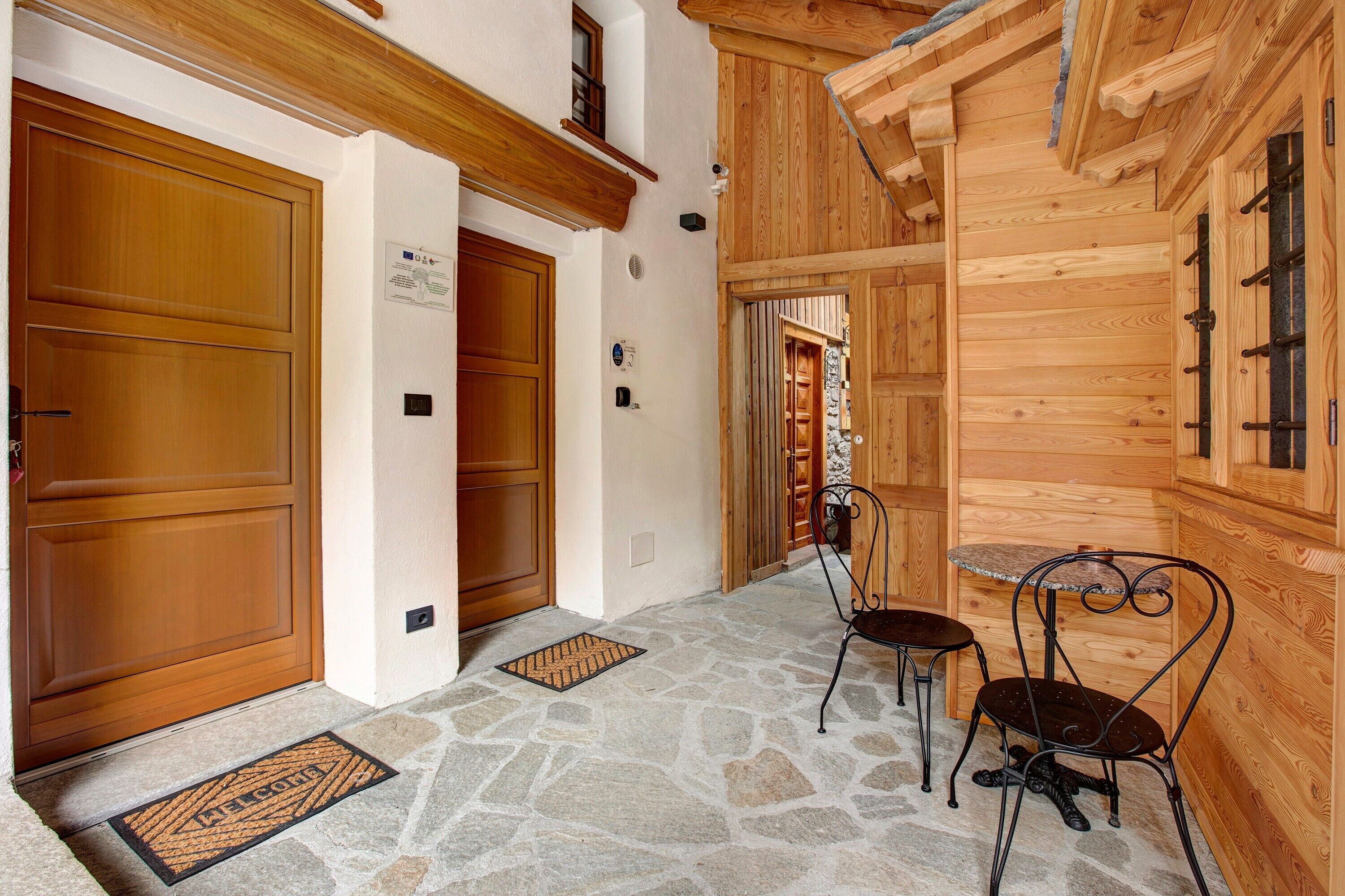 Un esterno in legno con pavimento in pietra, un tappetino d'ingresso e due sedie in metallo.