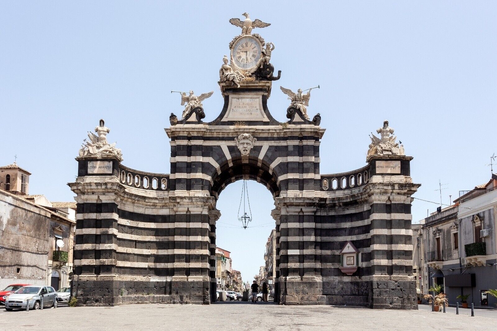 A historic stone archway with inscriptions and statues, leading to a street with parked cars and buildings.