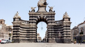 Un arc en pierre d’époque, orné d’inscriptions et de statues, qui mène à une rue bordée de voitures garées et d’immeubles.