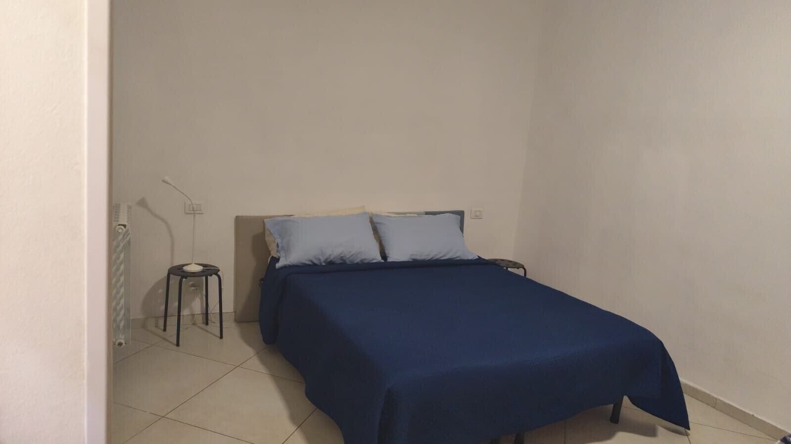 Une petite chambre avec un lit simple, une couverture bleue, une petite table et un radiateur.