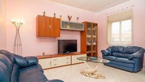 Een woonkamer met een televisie, houten kasten, een glazen salontafel, een blauwe bank en een witte kat op een deken.