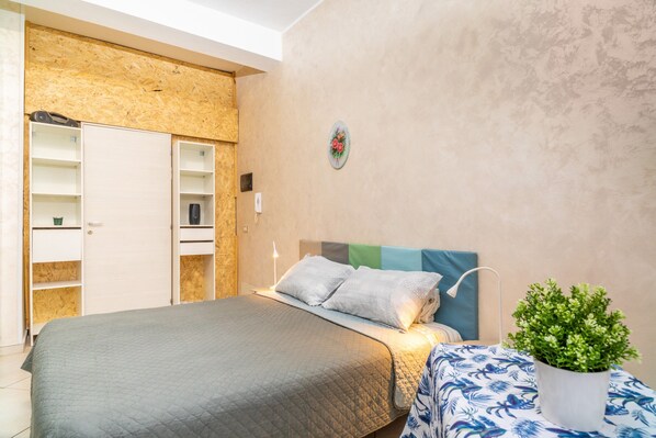 A bedroom with a bed, a nightstand, a potted plant, a wardrobe, and a wall-mounted shelf.