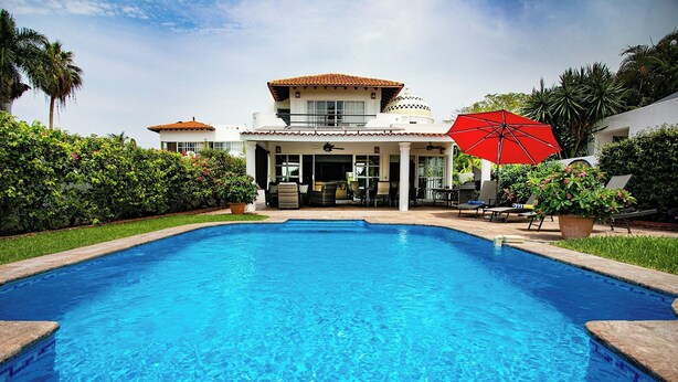 A swimming pool with a clear blue water surface, surrounded by a well-maintained garden and a building with a tiled roof. - La Puntilla, Luxury 3-bedroom, 3-bath home with heated pool in gated community (La Cruz de Huanacaxtle)