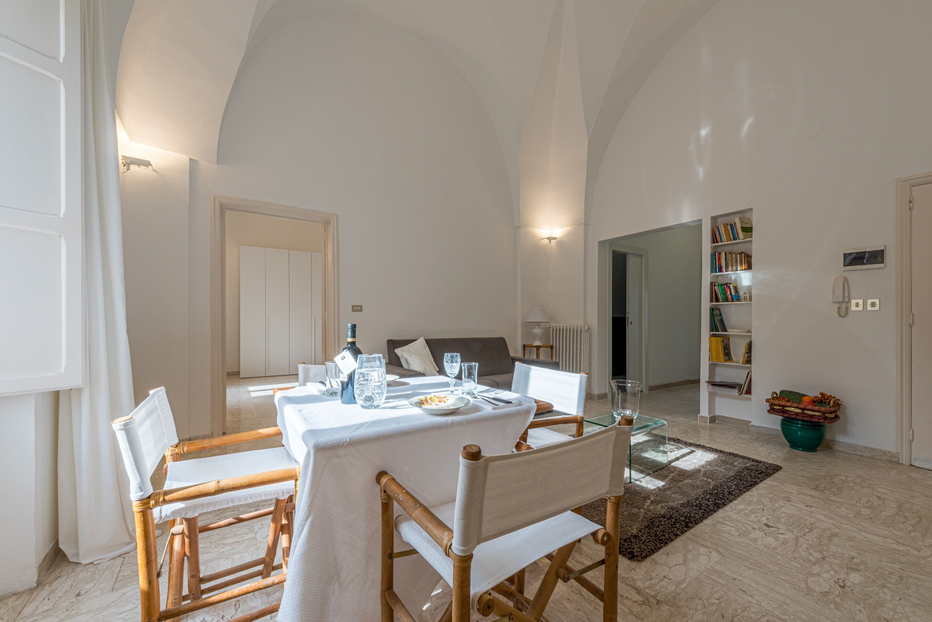 Un área de comedor con una mesa dispuesta para cuatro personas, un sofá al fondo y una estantería con libros.