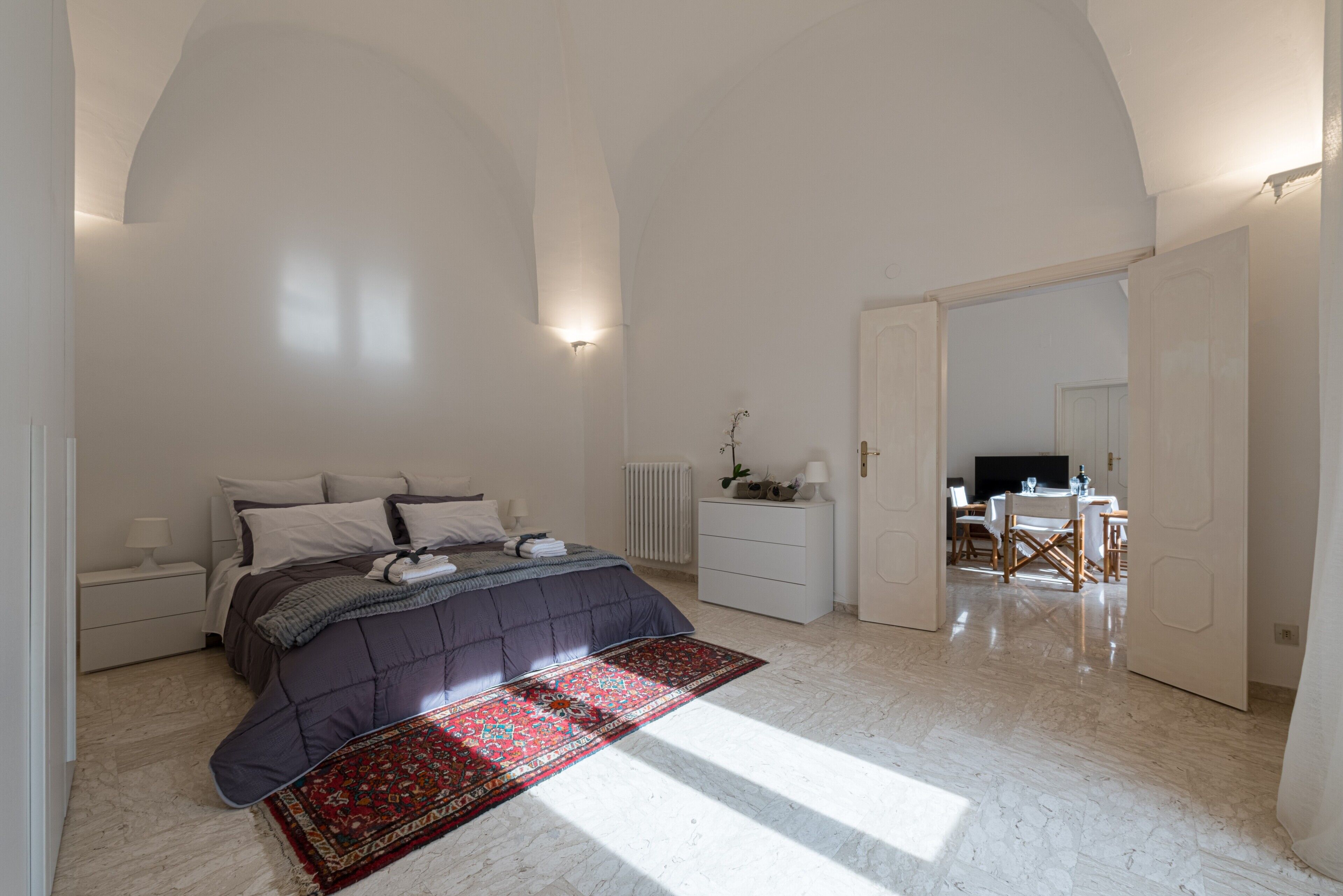 A bedroom with a bed, bedside tables, a dresser, a red patterned rug, and a dining area with chairs and a table.