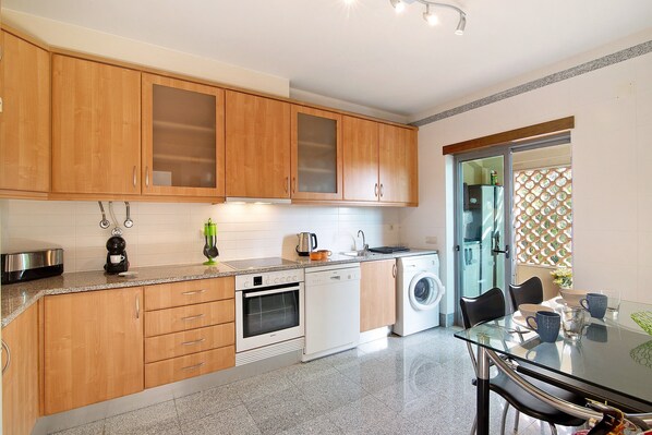 Una cocina moderna con armarios de madera, lavavajillas, horno y una mesa de vidrio con sillas.