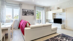 A modern living room with a white sofa, a dining table with chairs, a flat-screen TV, and a cityscape painting on the wall. - Villa 'Joel' with Sea View, Wi-Fi and Air Conditioning (Fontane Bianche)