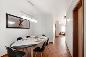 A dining area with a table set for four, chairs, a chandelier, a framed picture on the wall, and a view into another room.