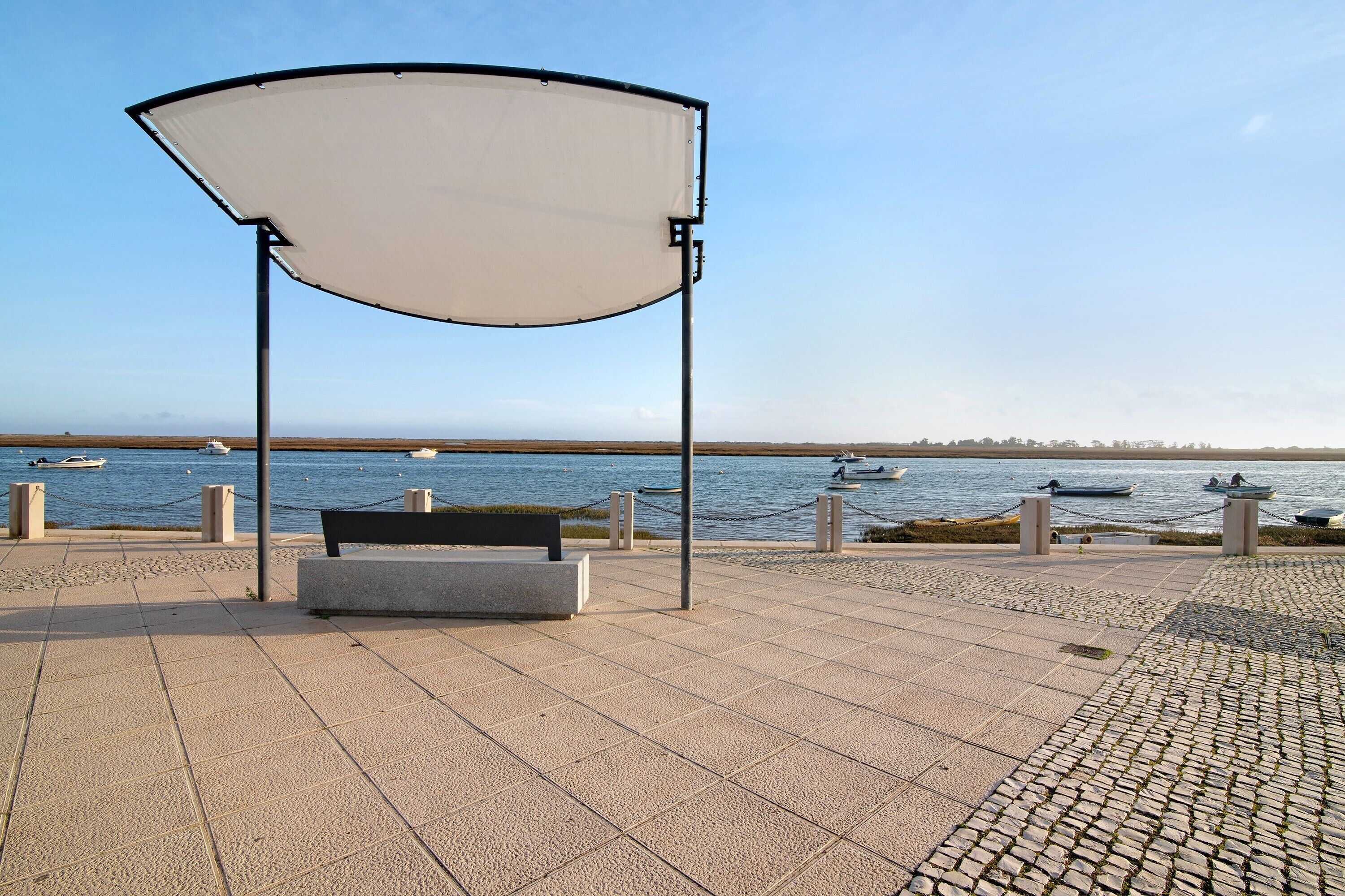 Un sendero pavimentado con un banco y una pérgola, con vistas a un cuerpo de agua con barcos.