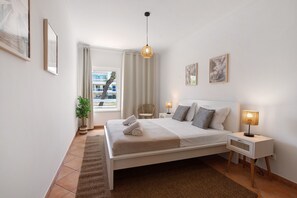 Une chambre à coucher comprenant un lit, des tables de chevet, un luminaire suspendu, une fenêtre avec des rideaux et des œuvres d’art accrochées au mur.